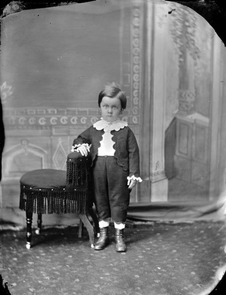 Studio portrait of an unidentified boy posing standing with his right arm resting on a chair.