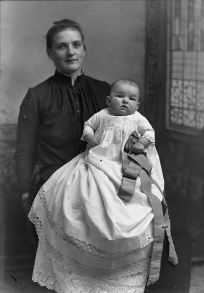 Studio portrait in front of a painted backdrop of an unidentified woman posing sitting and holding an infant.