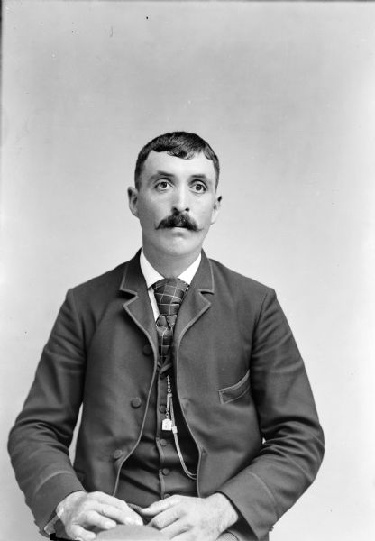 Waist-up studio portrait of an unidentified man with a moustache posing sitting. He is wearing a dark-colored suit coat, vest, necktie, and watch chain.