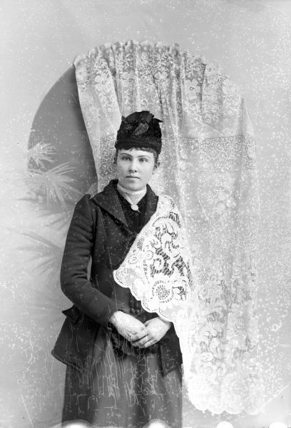 Studio portrait of an unidentified woman posing standing in front of a light-colored backdrop. She has a light-colored lace drapery over her left shoulder, and is wearing a dark-colored coat, skirt, blouse, and hat.
