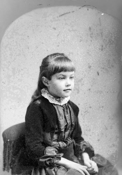 Studio portrait of an unidentified girl posing sitting. She is looking to the right and is wearing a dark-colored dress with a light-colored collar.