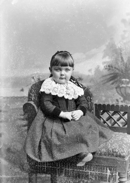 Studio portrait in front of a painted backdrop of an unidentified girl posing sitting. She is wearing a dark-colored dress with a light-colored collar.
