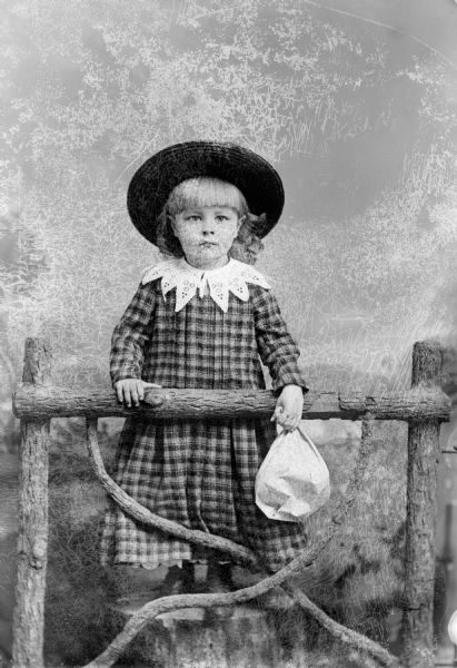 Studio portrait in front of a painted backdrop of an unidentified girl posing standing on a stump behind a prop wooden fence. She is holding a light-colored paper bag in her left hand, and is wearing a dark-colored plaid dress with a light-colored collar, and a hat.