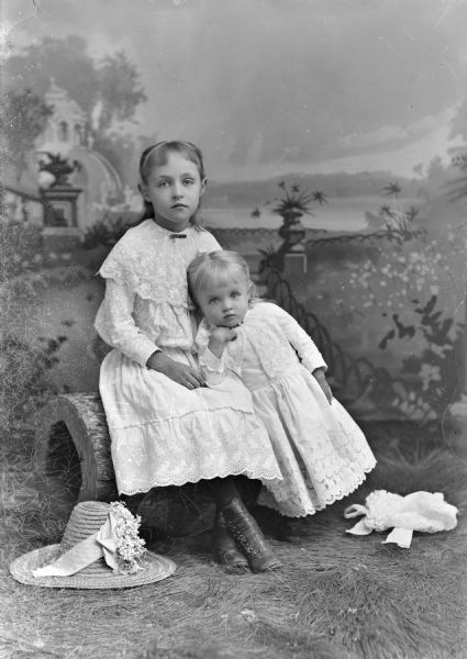 Studio portrait in front of a painted backdrop of two unidentified girls. They are both wearing light-colored dresses with lace trim. The older girl is sitting on a log on the left, and the younger girl is leaning on her lap on the right. There is a straw hat and a bonnet lying on the ground which is covered with fake grass.