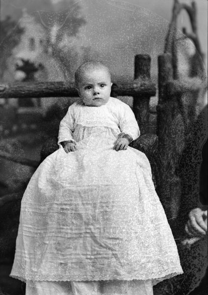 Studio portrait of an unidentified infant sitting in front of a prop wooden fence. The infant is wearing a long, light-colored dress. Another person is sitting off-camera on the right.