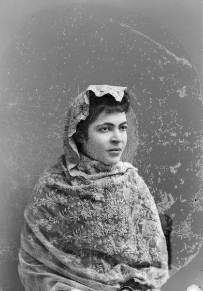 Waist-up studio portrait of an unidentified woman posing sitting. She is wearing a light-colored lace shawl, which is wrapped around her torso and over her head.