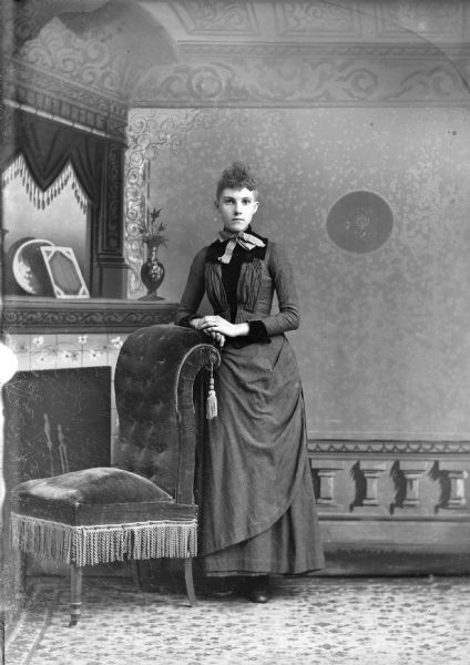 Full-length studio portrait in front of a painted backdrop of an unidentified woman posing standing with her right arm resting on the back of an upholstered chair. She is wearing a dark-colored dress and a ribbon bow.