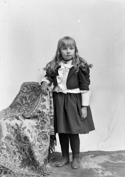 Full-length studio portrait of an unidentified girl with light-colored long hair posing standing with her right arm resting on the back of a chair draped with a cloth. She is wearing a dark-colored skirt, tights, short coat, and a light-colored blouse. In the background is a white backdrop.