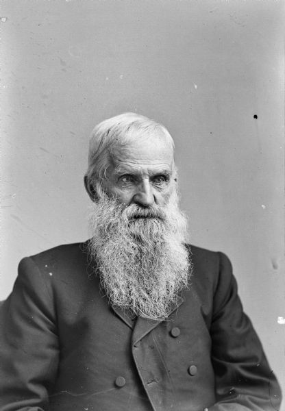 Waist-up studio portrait of an unidentified elderly man with a long beard posing sitting. He is wearing a dark-colored suit coat.