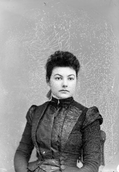 Waist-up studio portrait of an unidentified woman posing sitting. She is wearing a dark-colored dress and collar pin.
