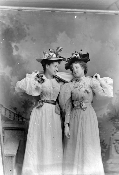 Three-quarter length studio portrait in front of a painted backdrop of two unidentified women posing standing while holding a single umbrella over their shoulders. Both women are wearing light-colored dresses and hats.