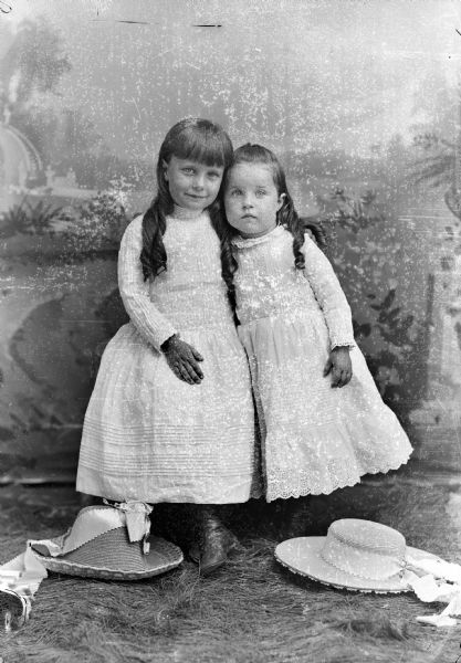 Full-length studio portrait in front of a painted backdrop of two unidentified girls posing while standing close together. Both of the girls are wearing light-colored dresses, and two hats are resting on the ground which is covered with fake grass.