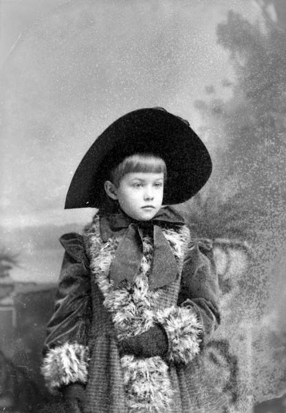 Waist-up studio portrait in front of a painted backdrop of an unidentified girl posing standing. She is wearing a dark-colored coat with fur trim, gloves, and a dark-colored hat tied around her neck with a large ribbon.