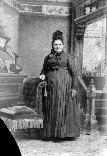 Full-length studio portrait in front of a painted backdrop of an elderly unidentified woman posing standing with her right hand on the back of an overstuffed chair. She is wearing a dark-colored dress and a dark-colored bonnet-type hat with long ribbons.
