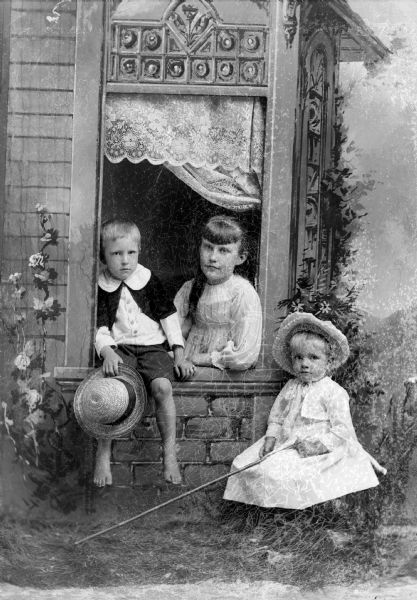 Studio portrait in front of a painted backdrop of three unidentified children posing around a prop window scene. There is a boy sitting on the window ledge with his legs dangling. He is holding a hat in his hand and is wearing dark-colored short pants and button-down light- and dark-colored shirt. There is a girl with long hair sitting behind the window in the center. She is wearing a light-colored dress. A small girl is sitting on the right and is holding a long pole. She is wearing a light-colored dress and a hat.