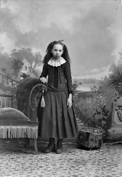 Full-length studio portrait in front of a painted backdrop of an unidentified girl posing standing, with her right hand on the back of an upholstered chair. She is wearing a dark-colored dress with a wide, light-colored lace collar.