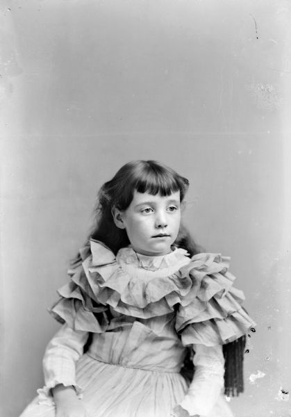 Waist-up studio portrait of an unidentified girl posing sitting in a chair with a tasseled back. She is wearing a light-colored dress with a ruffled bodice.
