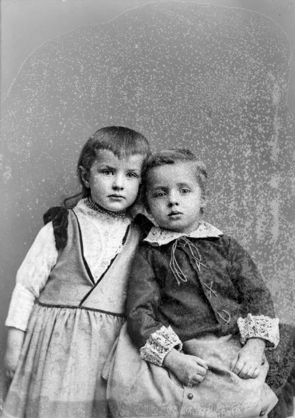 Three-quarter length studio portrait in front of a painted backdrop of two unidentified children. The girl standing on the left is wearing a light-colored dress and short necklace. The boy sitting on the right is wearing a light-colored skirt and dark-colored lace-up shirt with light-colored lace cuffs and collar.
