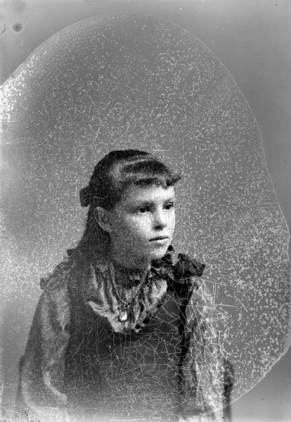 Waist-up studio portrait of an unidentified girl posing sitting. She is wearing a dark-colored dress and blouse, and a wide linked necklace with locket.