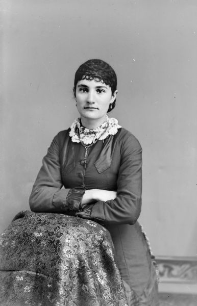 Three-quarter length sudio portrait of an unidentified woman posing standing with her arms resting on the back of an object draped with a cloth. She is wearing a dark-colored dress with a wide light-colored lace collar and a necklace with a cross.
