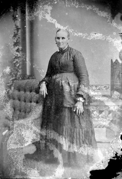 Copy photograph of a full-length studio portrait in front of a painted backdrop of an elderly unidentified woman posing standing with her right hand resting on the back of an upholstered chair. She is wearing a dark-colored dress and earrings.