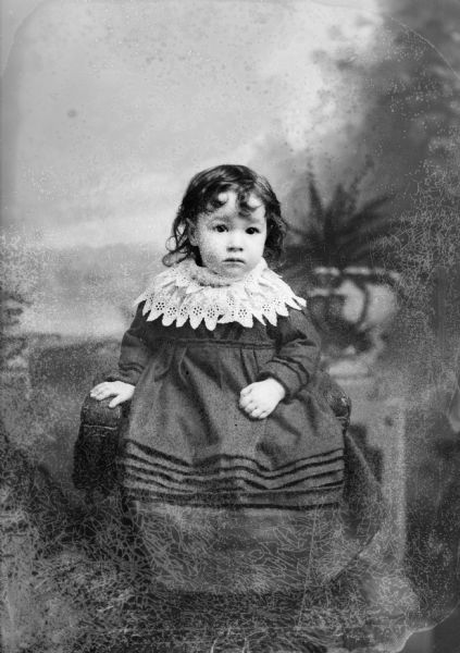 Full-length studio portrait in front of a painted backdrop of an unidentified girl posing sitting. She is wearing a dark-colored dress with a wide light-colored lace collar.
