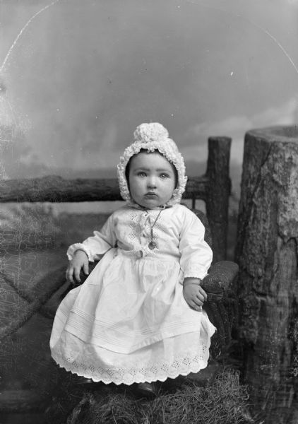 Studio portrait in front of a painted backdrop of an unidentified young child posing sitting and wearing a light-colored dress, bonnet, and a necklace with a locket. The child is sitting in a chair with her feet on fake grass. On the right is a tree stump, and in the background is a prop fence.