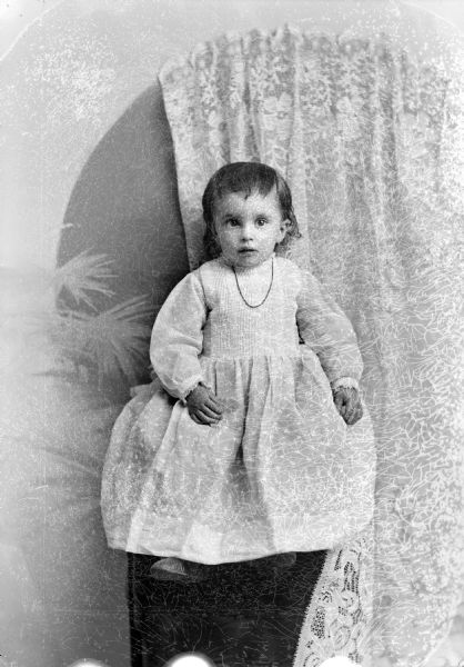 Studio portrait of an unidentified young child posing sitting on a raised platform. The child is wearing a light-colored dress and a necklace. Behind the child on the wall is a lace curtain and painted backdrop.