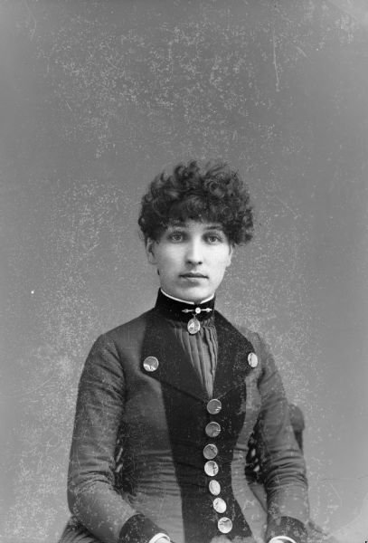 Waist-up studio portrait of an unidentified woman posing sitting. She is wearing a dark-colored dress with darker-colored trim, a button-down/pleated bodice, and a collar pin.