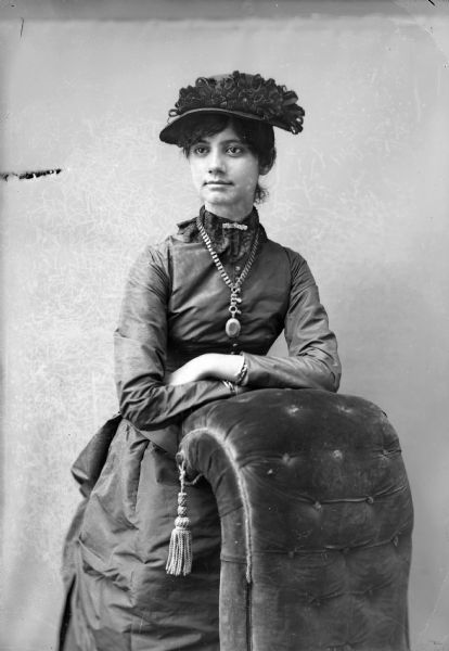 Three-quarter length studio portrait of an unidentified woman posing standing with her arms resting on the back of an overstuffed chair. She is wearing a dark-colored dress, collar pin, large-linked necklace with a locket, and a hat.
