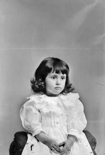 Waist-up studio portrait of an unidentified young girl posing sitting. She is wearing a light-colored dress and a necklace with a heart-shaped charm.