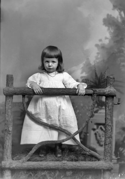 Full-length studio portrait in front of a painted backdrop of an unidentified young girl posing standing behind a wooden fence. She is wearing a light-colored dress and a necklace.