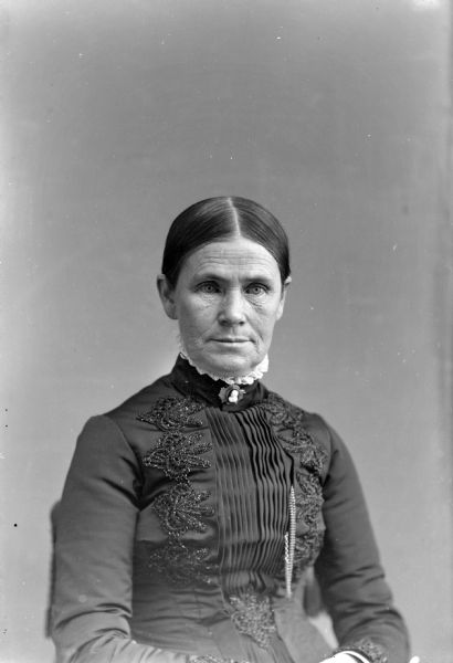 Waist-up studio portrait of an unidentified elderly woman posing sitting. She is wearing a dark-colored dress with a pleated bodice and an embroidered trim, a watch chain, and a cameo collar pin.