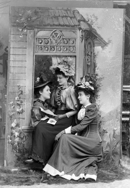 Studio portrait in front of a painted backdrop of three unidentified women. They are all wearing dark-colored dresses and hats and are posing around a prop window facade. The woman sitting in the center is holding a piece of paper and a photograph.