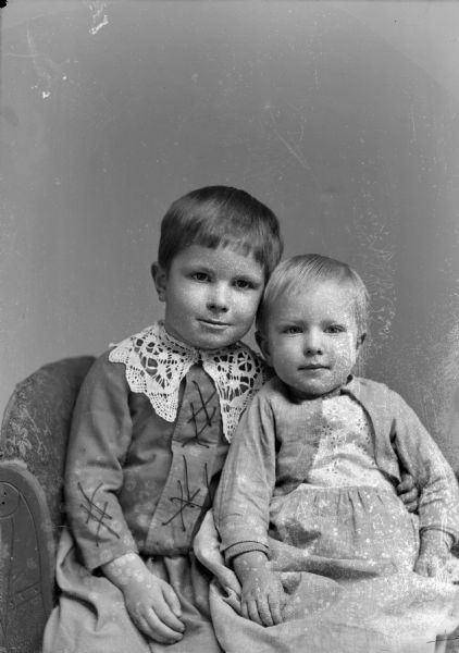 Studio portrait of an unidentified boy and younger child posing sitting. The boy sitting on the left is wearing a lace-up shirt with a wide light-colored lace collar. The child sitting on the right is wearing a light-colored dress.