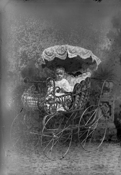 Studio portrait in front of a painted backdrop of an unidentified infant posing sitting in a baby carriage with an umbrella-type top. She is wearing a light-colored dress.
