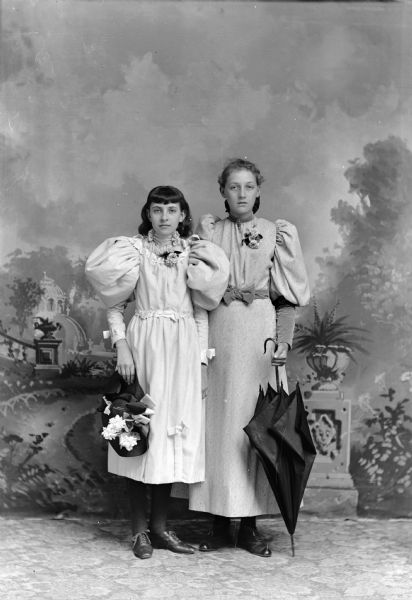 Full-length studio portrait in front of a painted backdrop of two unidentified European American women posing standing. The woman are wearing light-colored dresses and floral corsages. The shorter woman on the left is holding a hat in her right hand, and the taller woman on the right is holding an umbrella in her left hand.