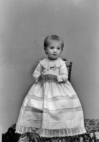 Studio portrait of an unidentified child posing sitting in a chair on top of a table covered with a cloth. She is wearing a light-colored dress.