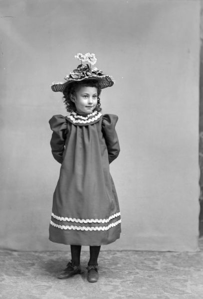 Full-length studio portrait of an unidentified girl posing standing with her hands behind her back. She is wearing a dark-colored dress with light-colored trim, a short necklace, and a hat with a floral and bow theme.
