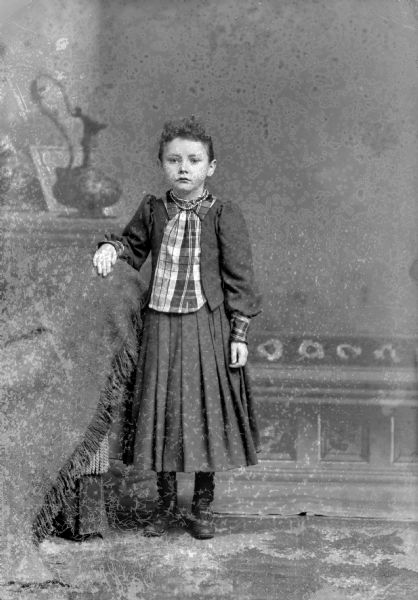 Full-length studio portrait in front of a painted backdrop of an unidentified girl posing standing with her right hand resting on the back of a chair draped with a cloth. She is wearing a dark-colored skirt and jacket, and a plaid blouse.