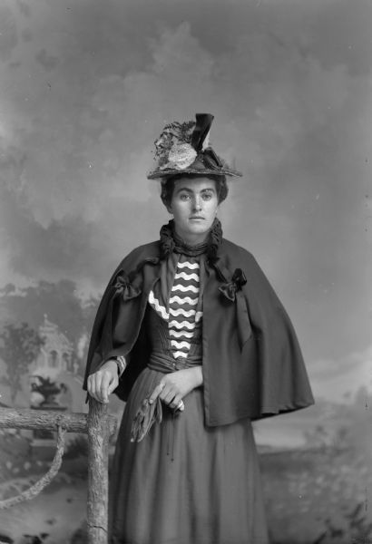 Three-quarter length studio portrait of an unidentified woman posing standing with her right hand resting on a prop wooden fence. She is holding a pouch-type purse in her left hand, and is wearing a dark-colored dress with a light-colored striped bodice, short dark-colored shawl, and floral hat with mesh veil.