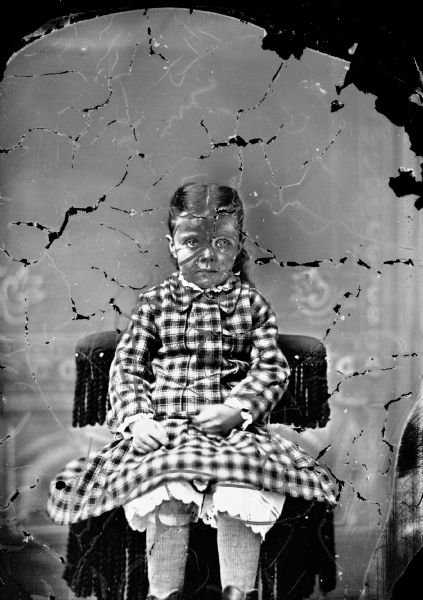 Studio portrait in front of a painted backdrop of an unidentified girl posing sitting in a chair with a tasseled back. She is wearing a plaid dress with light-colored knickers underneath.