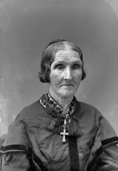 Quarter-length studio portrait of an unidentified elderly woman posing sitting. She is wearing a dark-colored tunic-style blouse with a lace collar, and a collar pin with a crucifix.