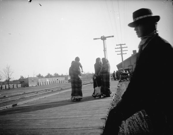 A Native American man is in the foreground, and three Native American women and two Native American children are standing on the wooden boardwalk near the depot. The stockyards are across the tracks in the background.