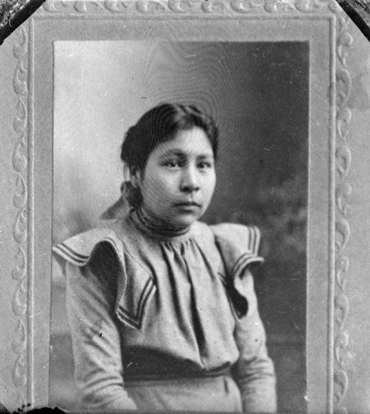 Copy photograph in front of a painted backdrop of a waist-up portrait of an unidentified young Native American woman posing sitting. She is wearing a dark-colored dress.