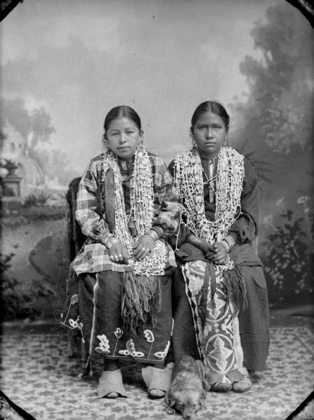 Studio portrait of two Native American girls posing sitting. They are wearing several long necklaces, dark-colored dresses, and earrings. A dog is lying on the ground between them.