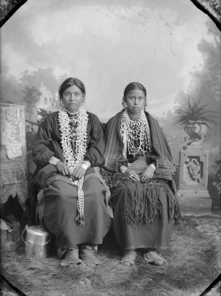 Full-length studio portrait in front of a painted backdrop of two Native American women posing sitting. Both woman are wearing several long necklaces, and are wrapped in fringed shawls. A bucket is sitting on the ground on the left.