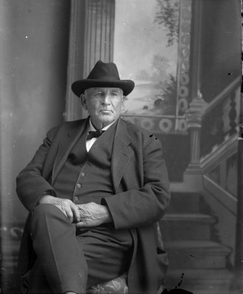 Studio portrait in front of a painted backdrop of an elderly European American man posing sitting. He is wearing a dark-colored suit coat, vest, bow tie, and hat. Identified as H.A. Bright.
