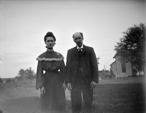 Outdoor portrait of a European American woman and man posing standing in a field. Behind them is a wooden building near trees in the background. The man is identified as Charlie Johnson, the son of Calvin Johnson.