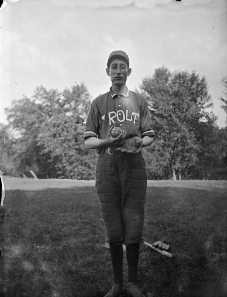 Outdoor portrait of a man posing and standing in a field of short grass. He is wearing a baseball uniform, with a jersey that reads "Trout" and is holding a baseball in one hand, and wearing a mitt in the other. A baseball bat and mitt are lying on the ground behind him. Identified as probably William Wensel.